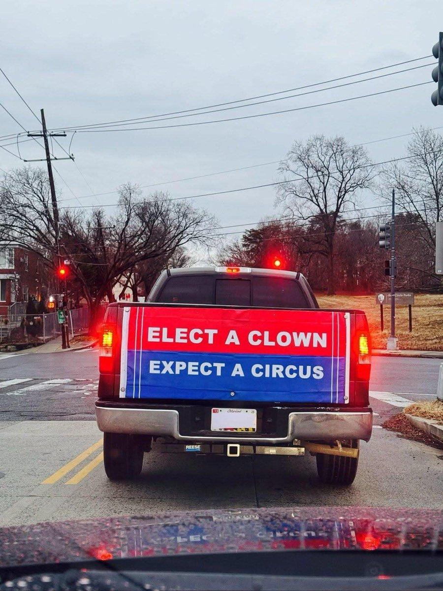 A pickup truck at a red light displays a large political banner attached to its tailgate. The banner has a bold red, white, and blue design resembling a campaign sign, with the words "ELECT A CLOWN - EXPECT A CIRCUS" written in capitalized white letters. The setting is an urban street with leafless trees, power lines, and traffic lights visible in the background under an overcast sky. The truck has a visible trailer hitch and a Maryland license plate, though the details of the plate are blurred. The image is taken from the perspective of another driver behind the truck.