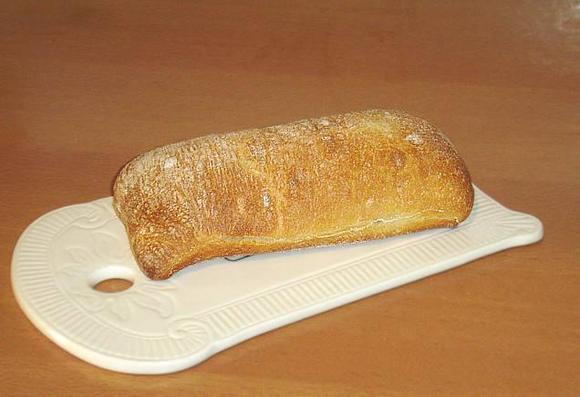 Ein Stück Ciabatta-Brot liegt auf einem weißen Schneidebrett mit einem rundem Griff. Das Brot hat eine längliche, flache Form und eine rustikale, unregelmäßige Oberfläche.