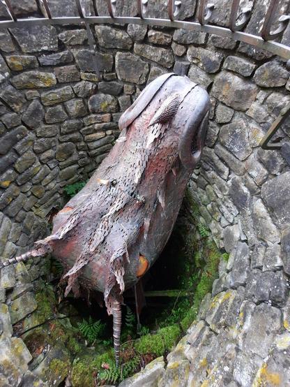 Photo of the inside of a stone well the sides of which are partially overgrown by moss and ferns. There is a large statue of a dragon head and neck made from metal peeking from the well with eyes made from large orange marbles.