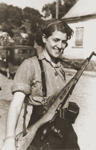 Photo of Sara Ginaite with her rifle standing in the street, a house and car in the background. 