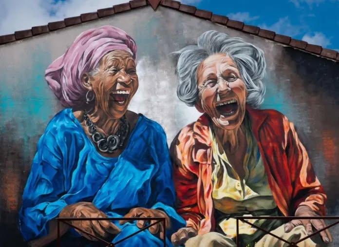 Mural of two elderly women laughing heartily, one in a blue robe with a pink headwrap, the other in orange and yellow clothes, painted on a rooftop wall in Éragny, France.