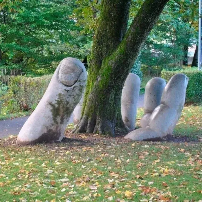 Sculpture of a giant stone hand rising from the ground, cupping a living tree in its palm, located in Glarus, Switzerland.