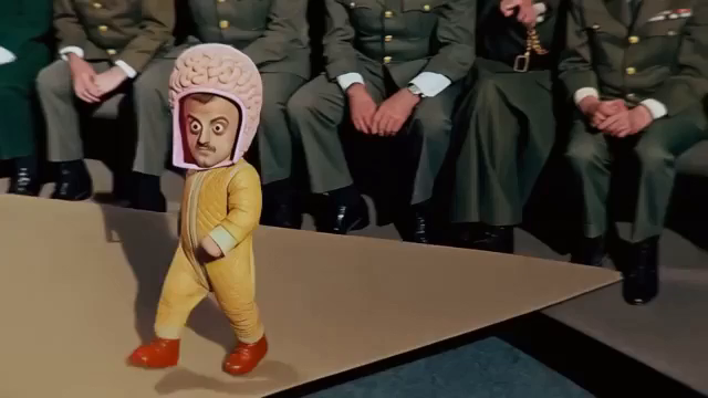 A clip from the music video for Igorrr's "ADHD." There's a little kid wearing a puffy mustard-colored suit and a big brain helmet, but he's not really a little kid, he's a shrunken man with a moustache. Then there's a giant brain float tethered to a car driving down a street. Next is a man lying on a couch in a psychologist's office looking uncomfortable and then the shrink himself appears. He looks like a shrink. Then there are some brains with 2, 3, or 4 long chicken legs walking on a beach. …