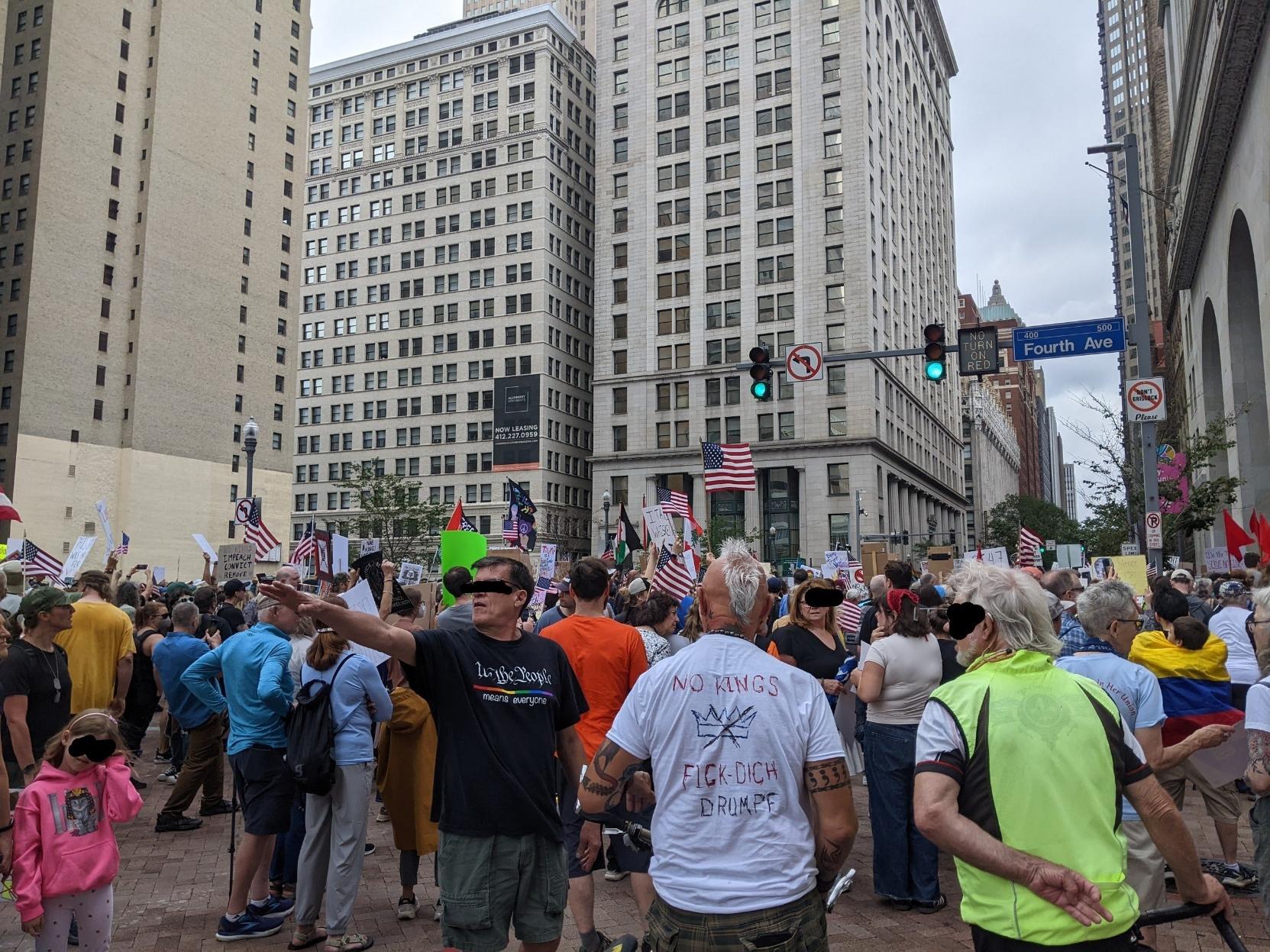 Crowd outside City county building, including little kid. The t shirt of a man with white mohawk style hair has "no kings"  and a crossed out crown in marker, and below that "Fick-Dich  Drumpf". Another man with his arm up has a dark blue T-shirt that says We the People in a US Constitution calligraphy font