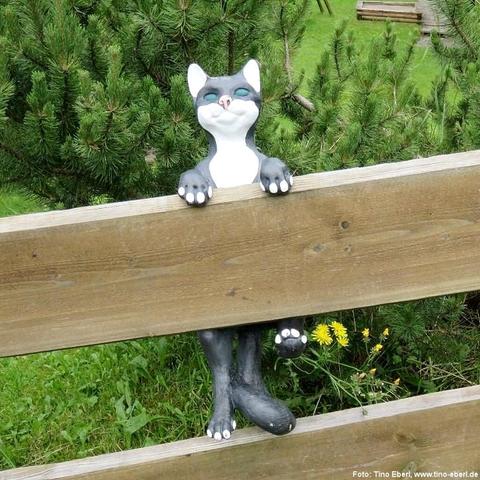 Katzenfigur aus glattem Kunststoff in dunklem Grauton mit weißer Brust und Gesichtspartie greift mit beiden Vorderpfoten über einen naturfarbenen Zaun; Hinterläufe und Schwanz ragen zwischen den Brettern hervor. Gesicht mit spitzen Ohren und in Blaugrün gehaltenen Augen blickt nach vorn. Hinter der Sitzfläche spannen sich dichte Zweige einer jungen Kiefer mit leuchtend grünen Nadeln, davor erscheint kurzgeschnittener Rasen mit vereinzelten gelben Löwenzahnblüten.
