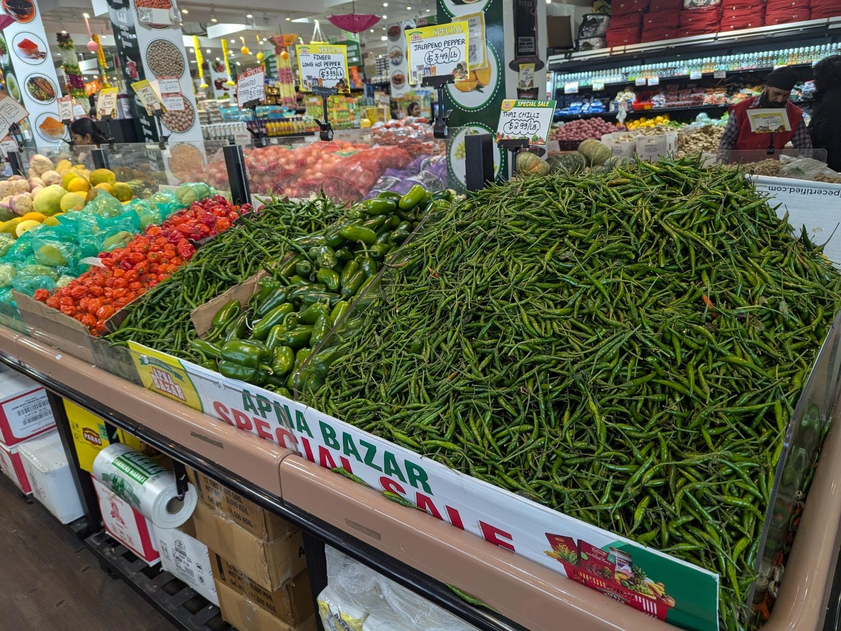 Grocery store with a stand/counter that has a pretty big mounds of green hot Thai chilis, jalapeños, long finger hot chillis, red habaneros
The text on a small plastic board shoring up the edge of the stand says Apna Bazar Special Sale