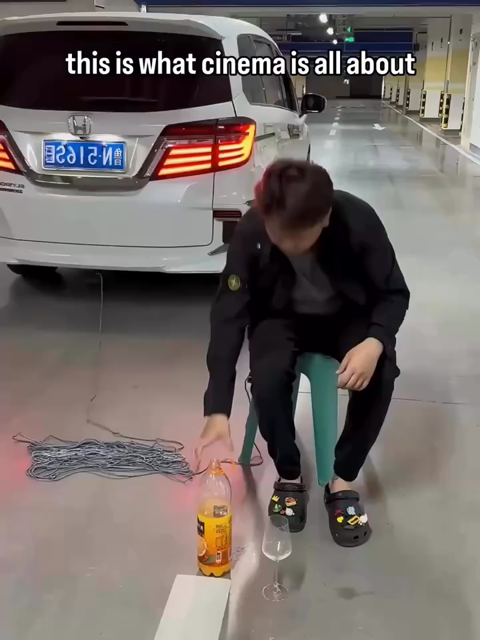 “This is what cinema is all about” over this video. In a parking garage, a person sits in a chair tied with rope to a car that’s pulling away. The person cuts the rope and pours themselves an orange drink. They drink the drink and start to enjoy it just as suddenly a scooter runs into them.