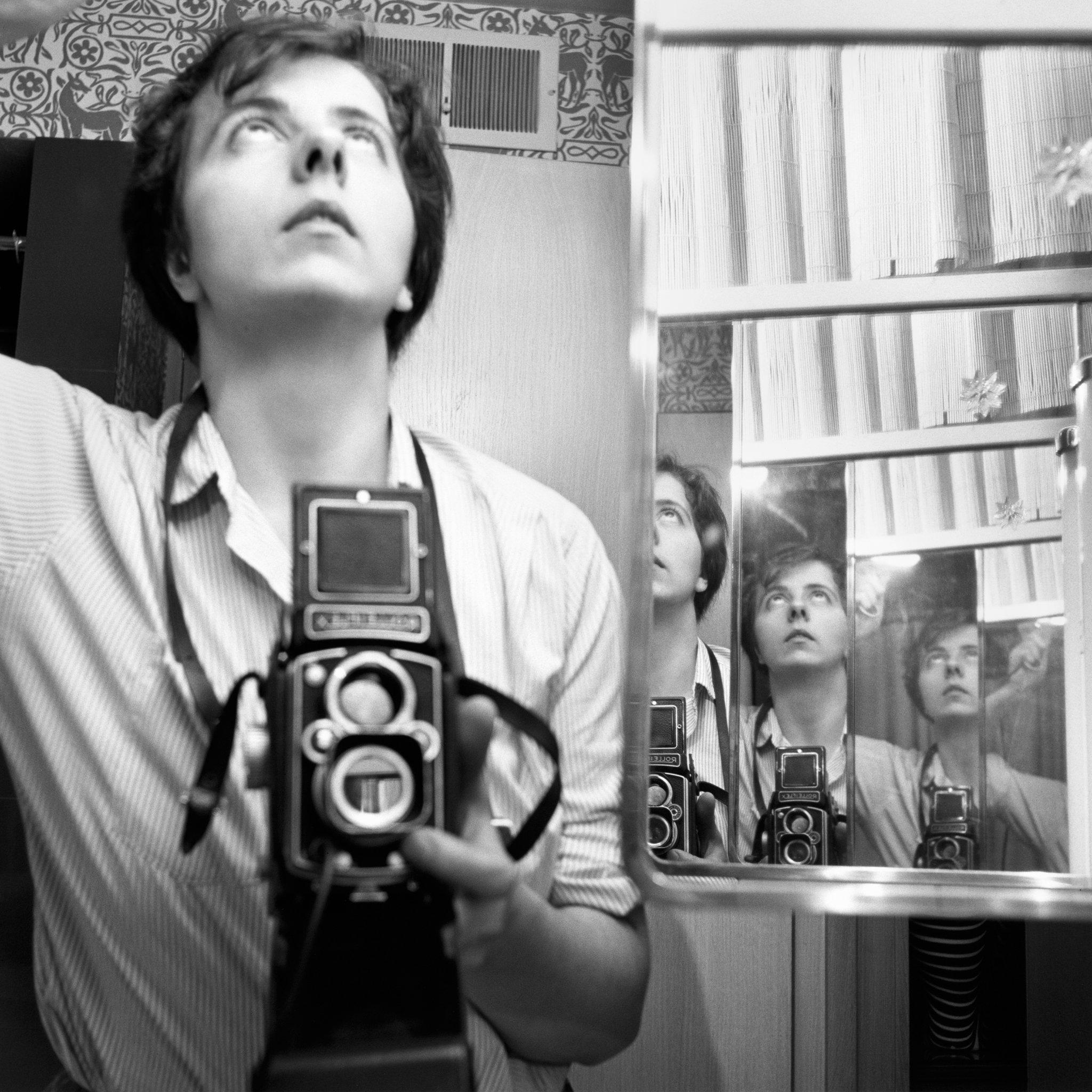 Black and white photo of a young white woman with short dark hair holding her camera below her neck and looking upward, reflected multiple times in large and small bathroom mirrors.