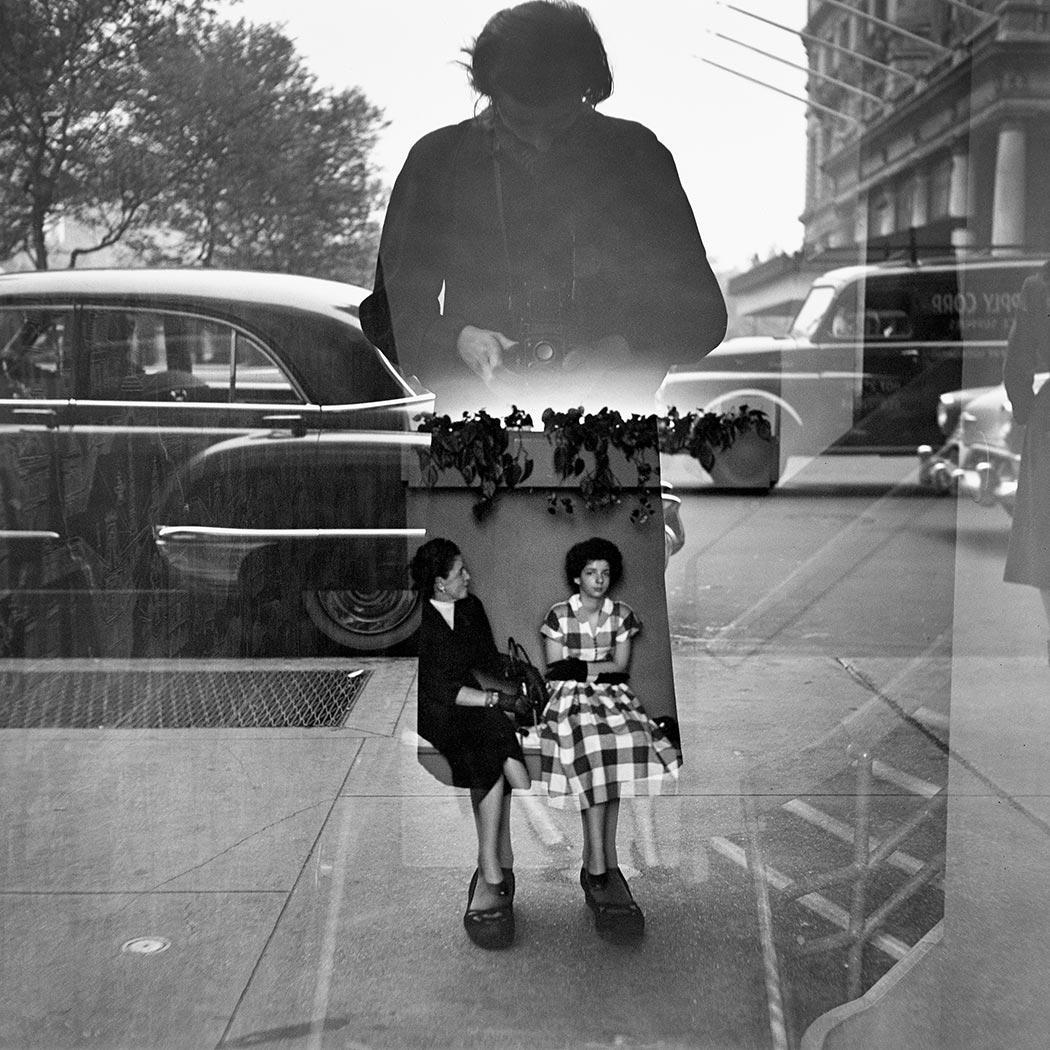 Black and white photo of a woman mostly in silhouette looking down at her camera and standing legs apart, reflected in a large exterior window showing a city street with cars behind her. Two women sitting inside on a bench are seen through her reflected silhouette.