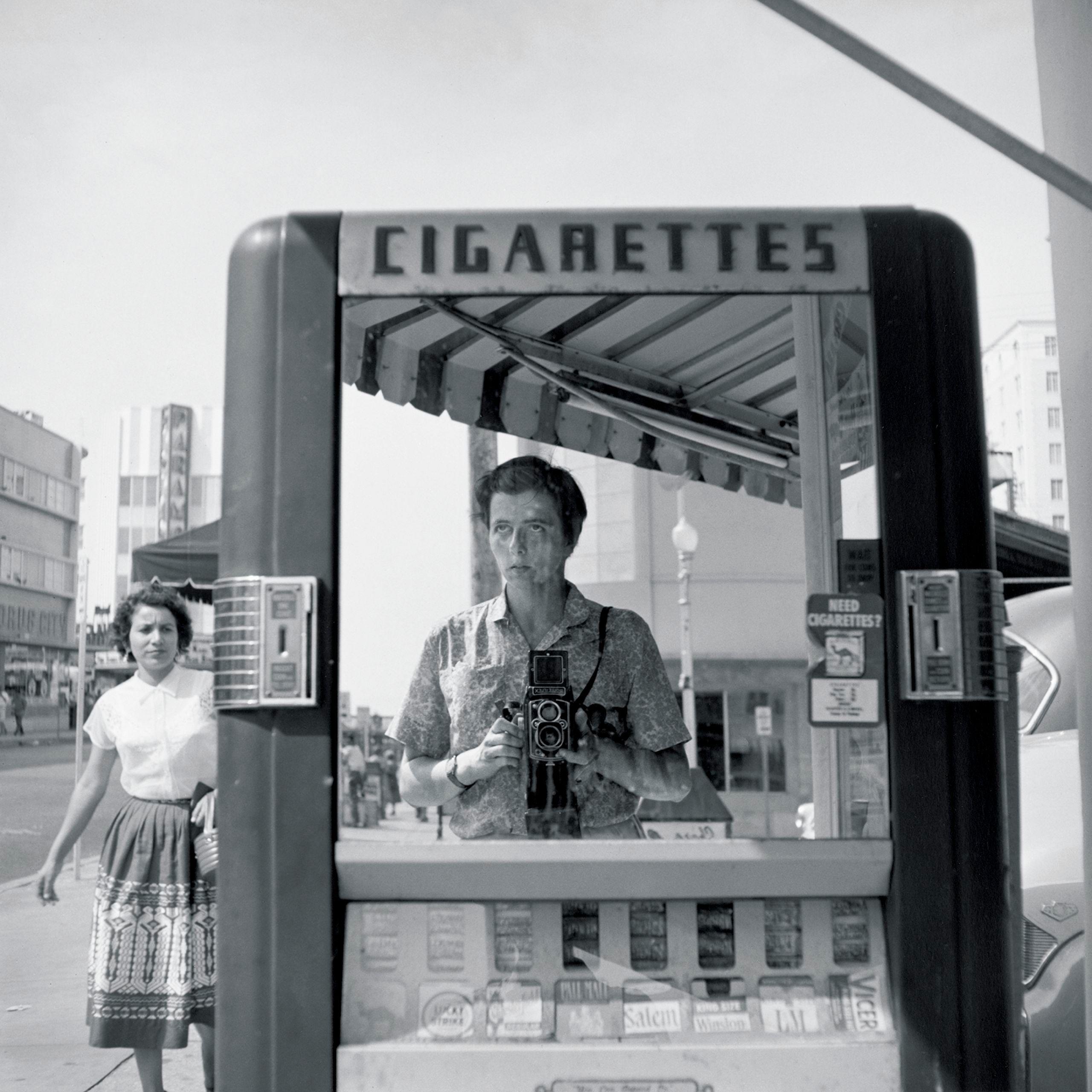 Black and white photo of a white woman with short dark hair holding her camera chest-level and looking outward, reflected in a cigarette machine on the street with a young woman walking on the sidewalk nearby.