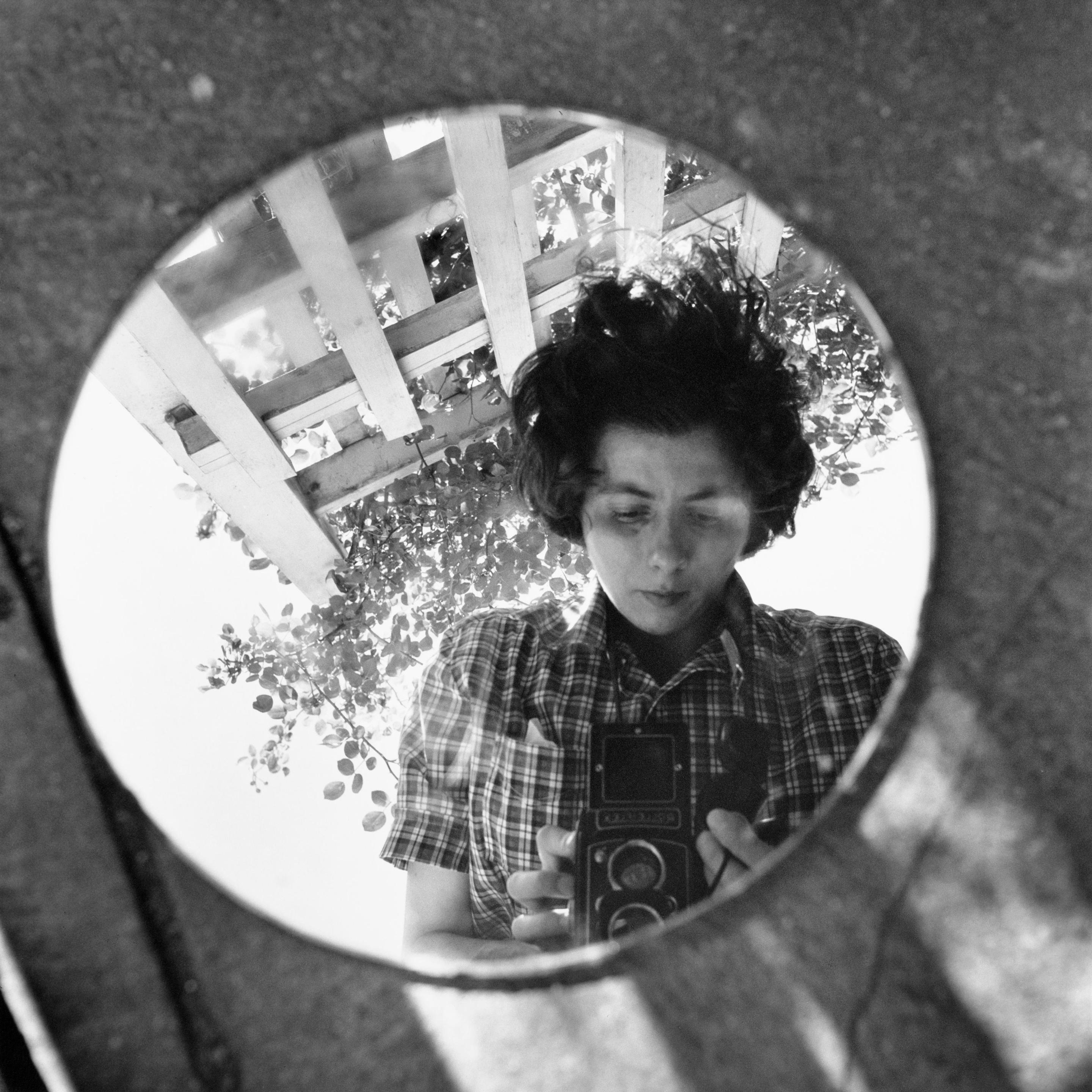 Black and white photo of a young white woman with short dark hair holding her camera and looking down, reflected in a round mirror on the ground, with a tall building and tree behind her.