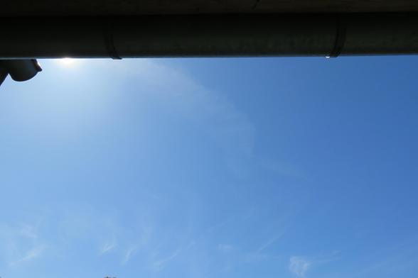 Im oberen Bereich des Bildes verläuft waagrecht ein Teil eines Dachvorsprungs mit einer Dachrinne. Dahinter ist ein strahlend blauer Himmel zu sehen, in dem sich eine große, helle Federwolke ausbreitet. Die Sonne scheint knapp hinter dem Dachvorsprung und erzeugt einen leuchtenden Schein. Am unteren Bildrand sind dünne, zarte Schleierwolken erkennbar.