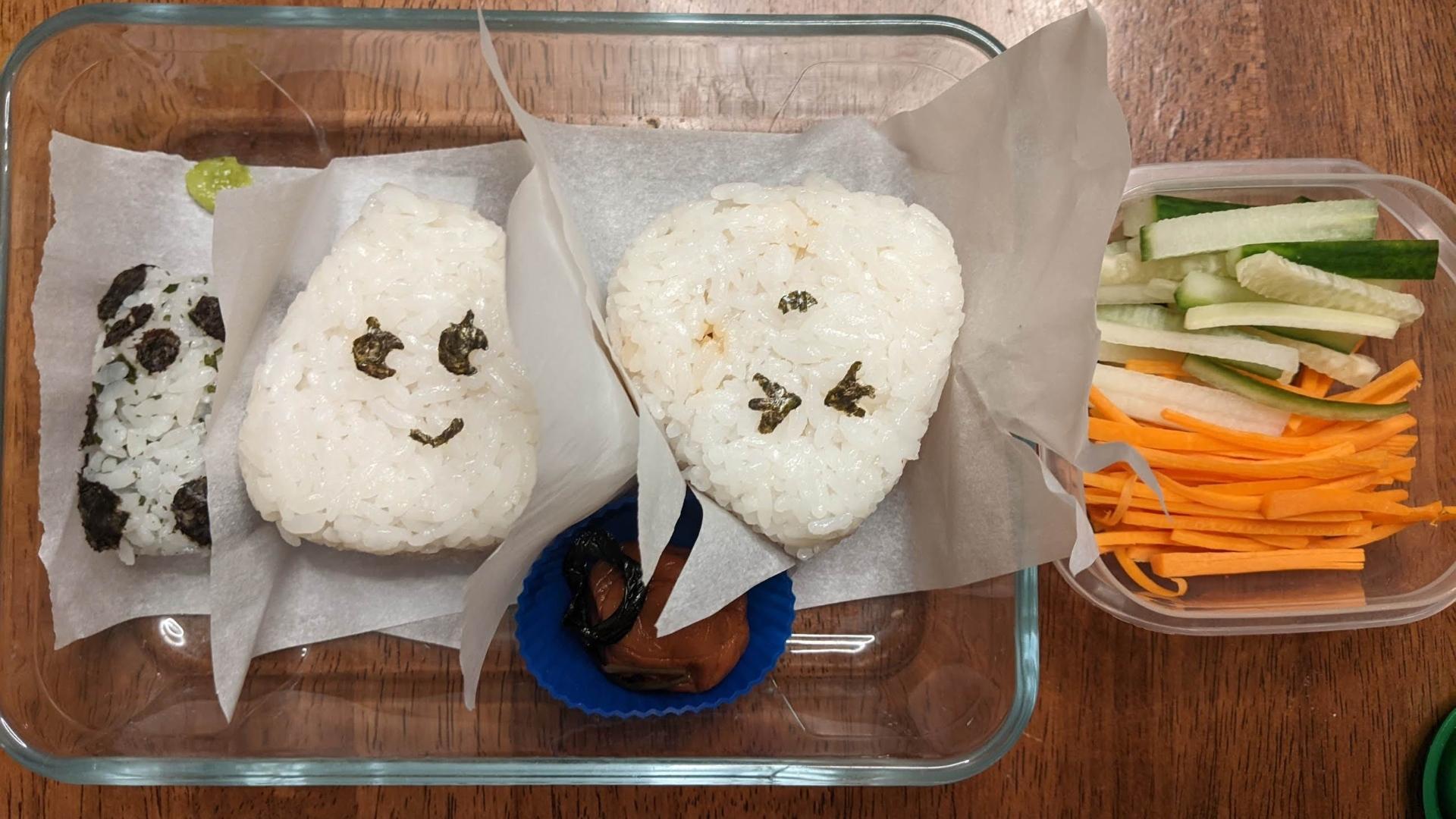 Rectangular glass container with a panda and also 2 triangular onigiri all on small pieces of parchment paper. The onigiri have eyes and a thin mouth with nori. A small round blue silicone holder has a pickled plum and shiso leaf in it. Another container to the right has thick matchstick size cut cucumbers and carrots
