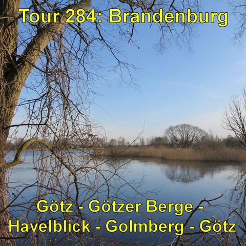 Ein ruhiger Flusslauf im Frühling. Die Bäume tragen keine Blätter. Die kahlen Äste spiegeln sich im stillen Wasser. Am Ufer stehen Schilf und niedrigere Vegetation. Der Himmel ist teilweise bewölkt, aber in einigen Bereichen blau und klar. Das Licht ist mild an einem sonnigen, aber kühlen Tag. Die Perspektive ist vom Ufer aus, mit Blick entlang des Flusses, umrahmt von den kahlen Ästen zweier großer Bäume im Vordergrund.