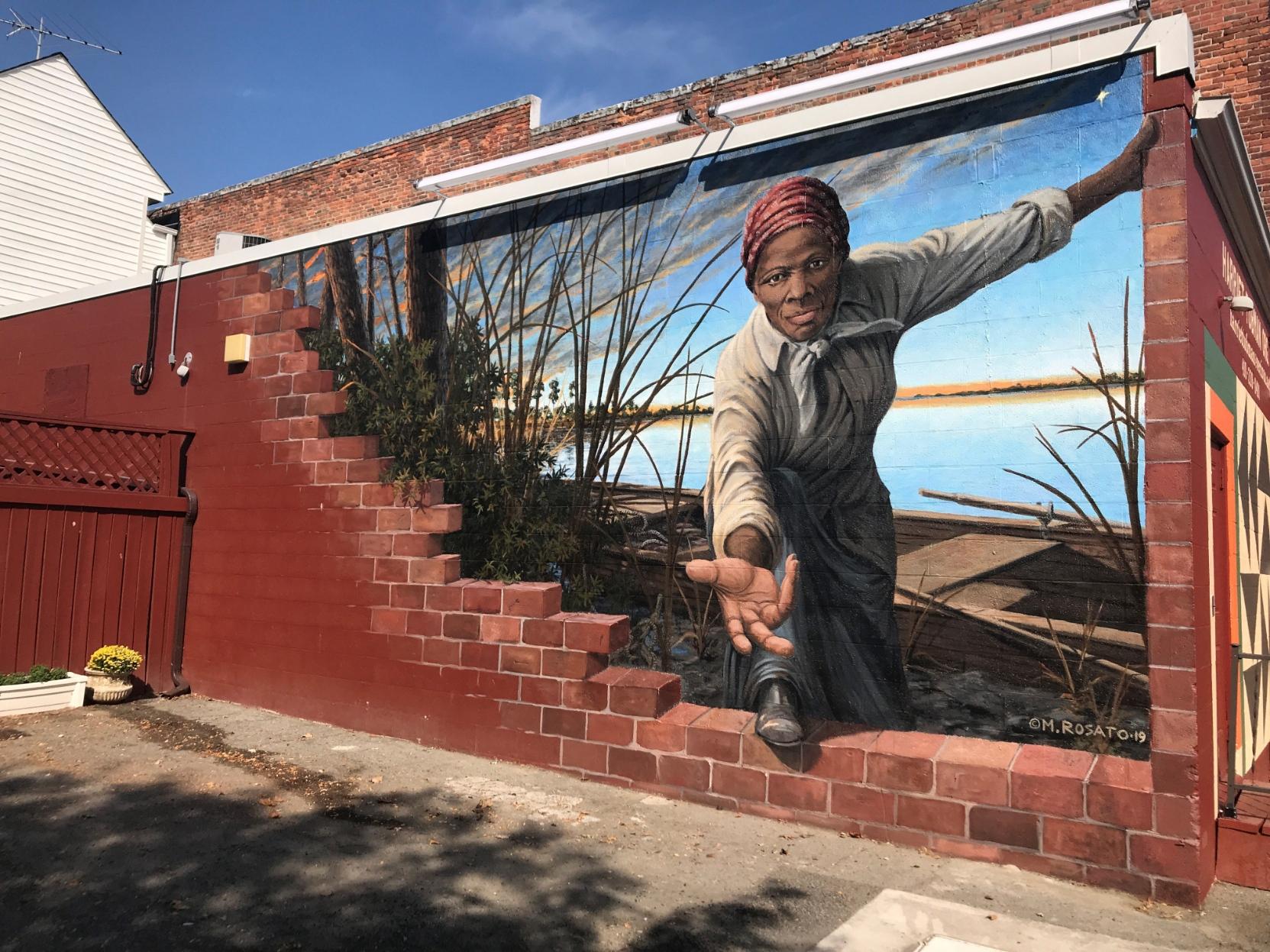 A mural by Michael Rosato on the side of a brick building next to a parking lot. The painting shows Tubman larger than life with blue skies and a river bank behind her, reaching through the wall she's broken through to offer a welcoming hand to those who would step through to freedom.
A mural on brick wall that looks as if the wall is partially crumbled, revealing a black woman reaching through to assist the viewer through the wall and onto a waterway with a rowboat.
https://harriettubmanmural.com/
Harriet Tubman mural artist Michael Rosato A mural by Michael Rosato on the side of a brick building next to a parking lot. The painting shows Tubman larger than life with blue skies and a river bank behind her, reaching through the wall she's broken through to offer a welcoming hand to those who would step through to freedom.
A mural on brick wall that looks as if the wall is partially crumbled, revealing a black woman reaching through to assist the viewer through the wall and onto a waterway with a rowboat.
https://harriettubmanmural.com/
Harriet Tubman mural artist Michael Rosato