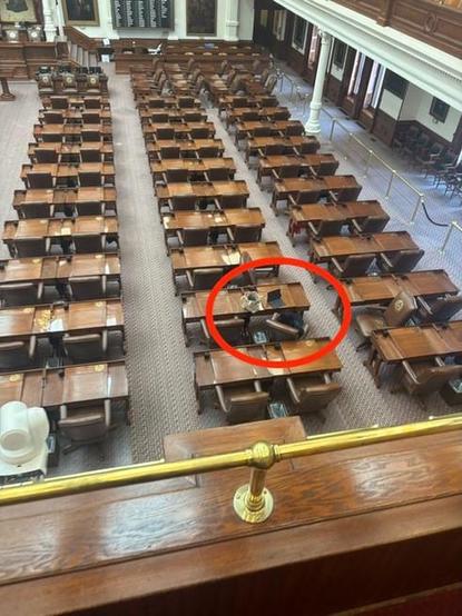 An overhead view of a legislative chamber with rows of wooden desks; one desk near the center is circled in red and is occupied.