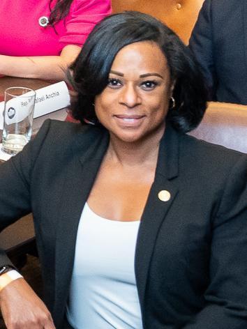 Rep. Nicole Collier wearing a black blazer and white top, smiling at the camera, with a nameplate and a glass of water visible in the background.