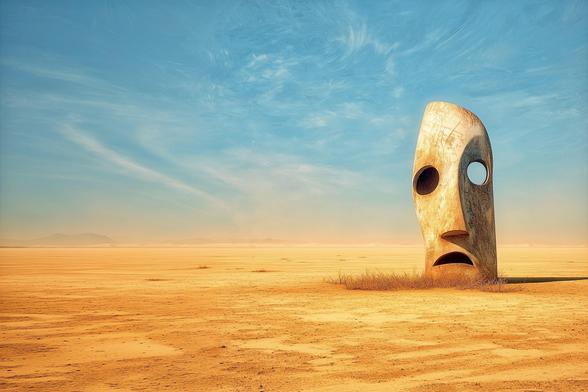A large, abstract stone sculpture resembling a stylized face with hollow eyes and an open mouth, standing alone in a vast desert under a blue sky.