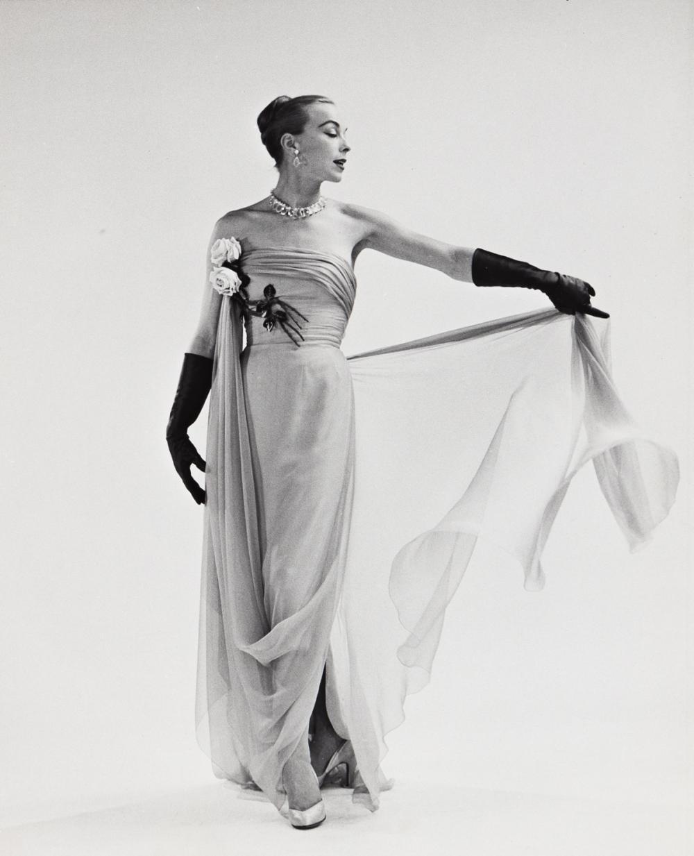Black and white photo of a slender white woman posing in blank space, wearing a light, long gown in a gauzy fabric, with a long swath draped from the waist down to her ankle and pulled back up to her chest, pinned with flowers. She wears dark gloves and holds some of the gauzy fabric out at her side so it sways back and forth.