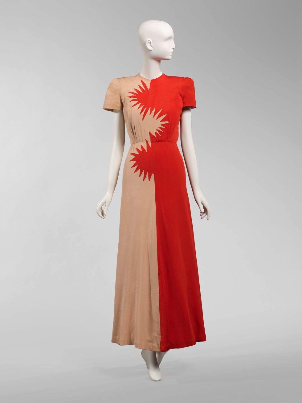 Color photo of a white mannequin wearing a dress with cap sleeves and a long skirt to the ankles. It is split vertically into beige and coral colors, with a starburst design in the center where the colors meet.