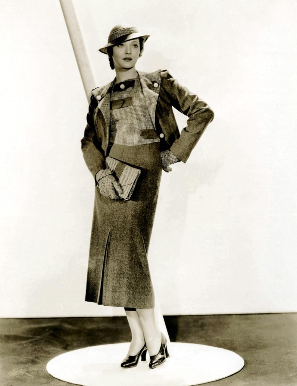 Black and white photo of a Sylvia Sidney posing in a room with dark floor and white wall, wearing a wool suit with two contrasting fabrics, one dark and one light with a subtle check pattern. The skirt has a slight flare and ends mid-calf. The jacked has large white buttons pinning back large lapels. She wears short gloves and a hat with a curving brim, and holds a small matching handbag.