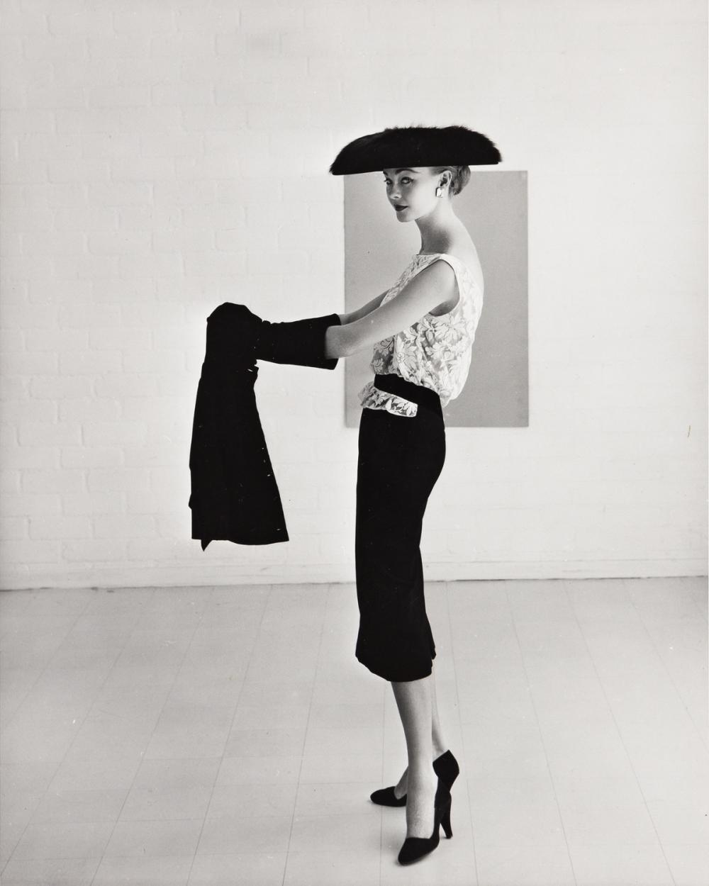 Black and white photo of a slender white woman posing in an empty room, wearing a dark hat shaped like a half-oval, a light lace sleeveless blouse, a dark pencil skirt, dark heels, and dark gloves.