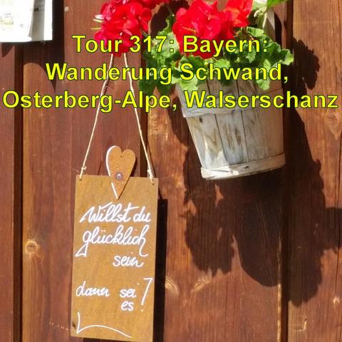 An der dunklen Holzwand einer Almhütte hängt ein dekoratives Schild mit einer Botschaft Willst du glücklich sein, dann asei es!, die mit einem Herzen verziert ist. Daneben befindet sich ein hängender Blumentopf mit leuchtend roten Blumen. Das Schild und der Blumentopf werfen ihre Schatten auf die Holzwand, was auf sonniges Wetter hindeutet. Die Szene vermittelt eine gemütliche, einladende Atmosphäre, wie sie oft in ländlichen oder alpinen Gebieten zu finden ist.