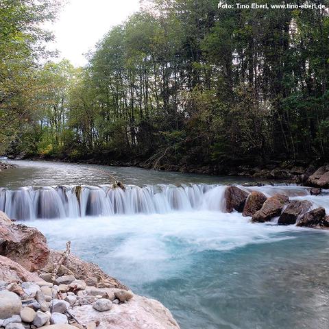 Ein klarer, türkisfarbener Fluss fließt durch eine bewaldete Landschaft. Im Vordergrund stürzt das Wasser über eine flache Felsstufe und bildet einen kleinen Wasserfall mit schäumender Gischt. Auf der linken Seite liegen größere und kleinere Steine am Ufer. Der Fluss ist von dichtem Wald gesäumt, dessen Bäume teils grüne, teils herbstlich verfärbte Blätter tragen. Das Wasser wirkt ruhig oberhalb des Wasserfalls und dynamisch darunter.