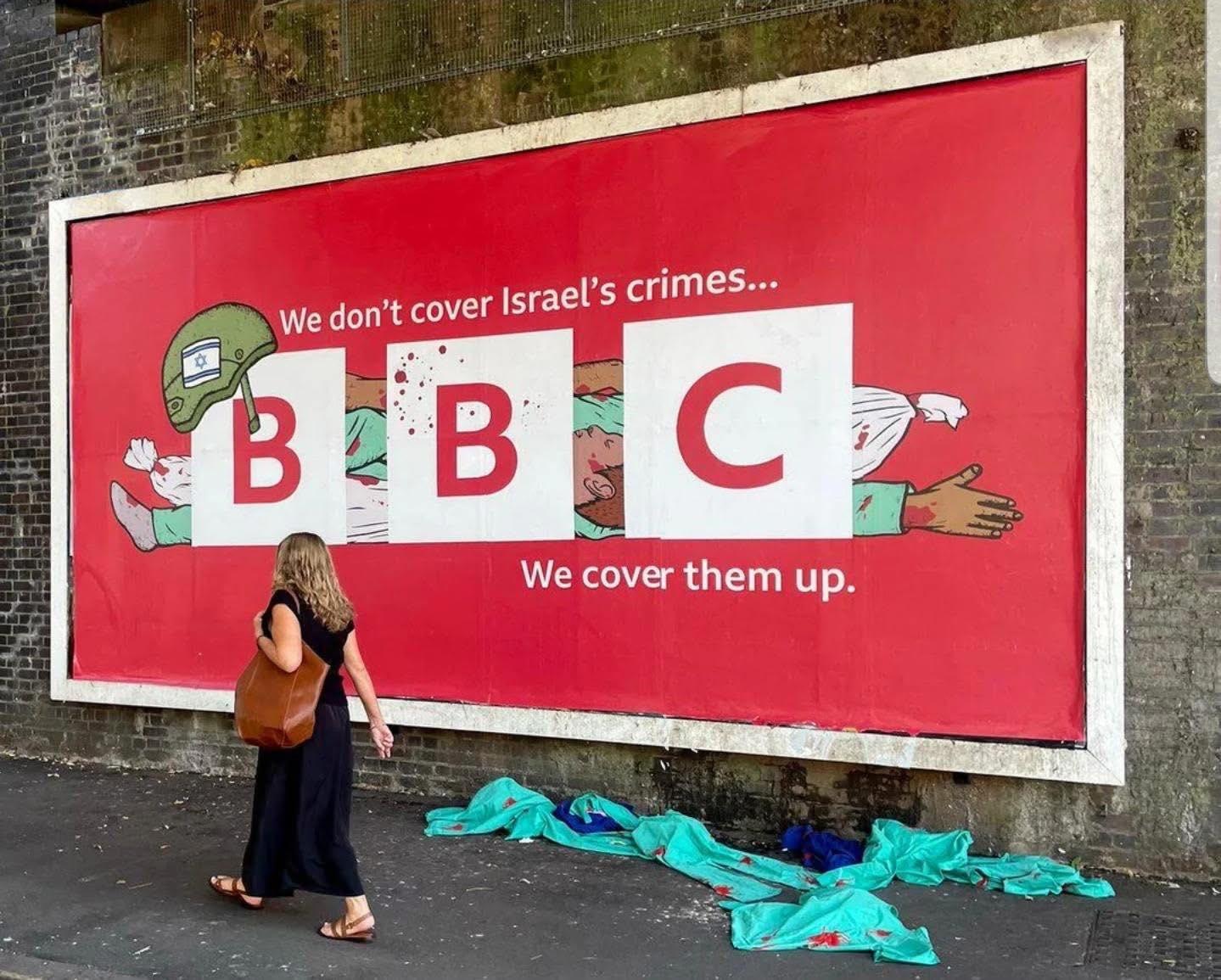 Political street art poster showing a critical message about BBC’s coverage of Israel. The artwork has a bright red background with the BBC logo in large white squares. Behind the letters, an illustration of a wounded body is partially covered by the blocks, suggesting censorship. A green helmet with the Israeli flag is drawn above the first "B". The text reads: “We don’t cover Israel’s crimes… We cover them up.” A woman in a black dress with a brown bag walks past the mural on the sidewalk. Political street art poster showing a critical message about BBC’s coverage of Israel. The artwork has a bright red background with the BBC logo in large white squares. Behind the letters, an illustration of a wounded body is partially covered by the blocks, suggesting censorship. A green helmet with the Israeli flag is drawn above the first "B". The text reads: “We don’t cover Israel’s crimes… We cover them up.” A woman in a black dress with a brown bag walks past the mural on the sidewalk.