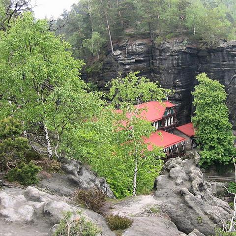Zwischen moosbedeckten Felsen und grünem Mischwald ragt ein Gebäude mit mehreren roten Dächern hervor. Es ist teilweise von Bäumen verdeckt und steht dicht an einer hohen, dunklen Felswand. Die Architektur weist auf ein größeres Haus mit Holzverkleidung und Fenstern über mehrere Etagen hin. Im Vordergrund befinden sich helle Sandsteinfelsen und niedrige Vegetation. Die Umgebung ist felsig und bewaldet mit Birken und Nadelbäumen.