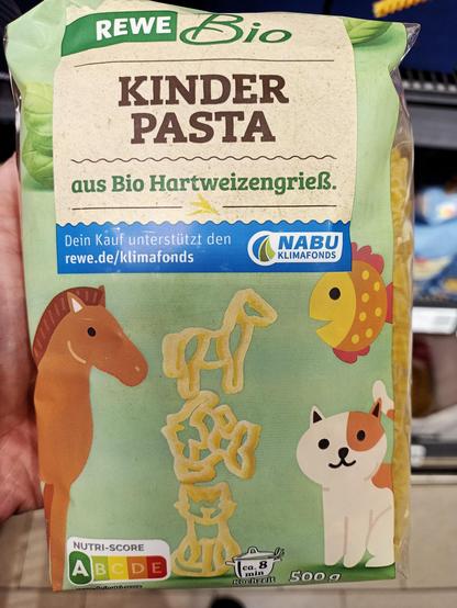 Die Verpackung zeigt eine grüne Tüte „REWE Bio Kinder Pasta“ aus Bio-Hartweizengrieß. Auf der Vorderseite sind Tierformen aus Pasta abgebildet (z. B. Giraffe, Löwe), daneben bunte Zeichnungen eines Pferdes, einer Katze und eines Fisches. Unten links ist der Nutri-Score A, rechts die Kochzeit (ca. 8 Minuten) und das Gewicht (500 g) angegeben. Ein blauer Hinweis informiert, dass der Kauf den NABU Klimafonds unterstützt.
