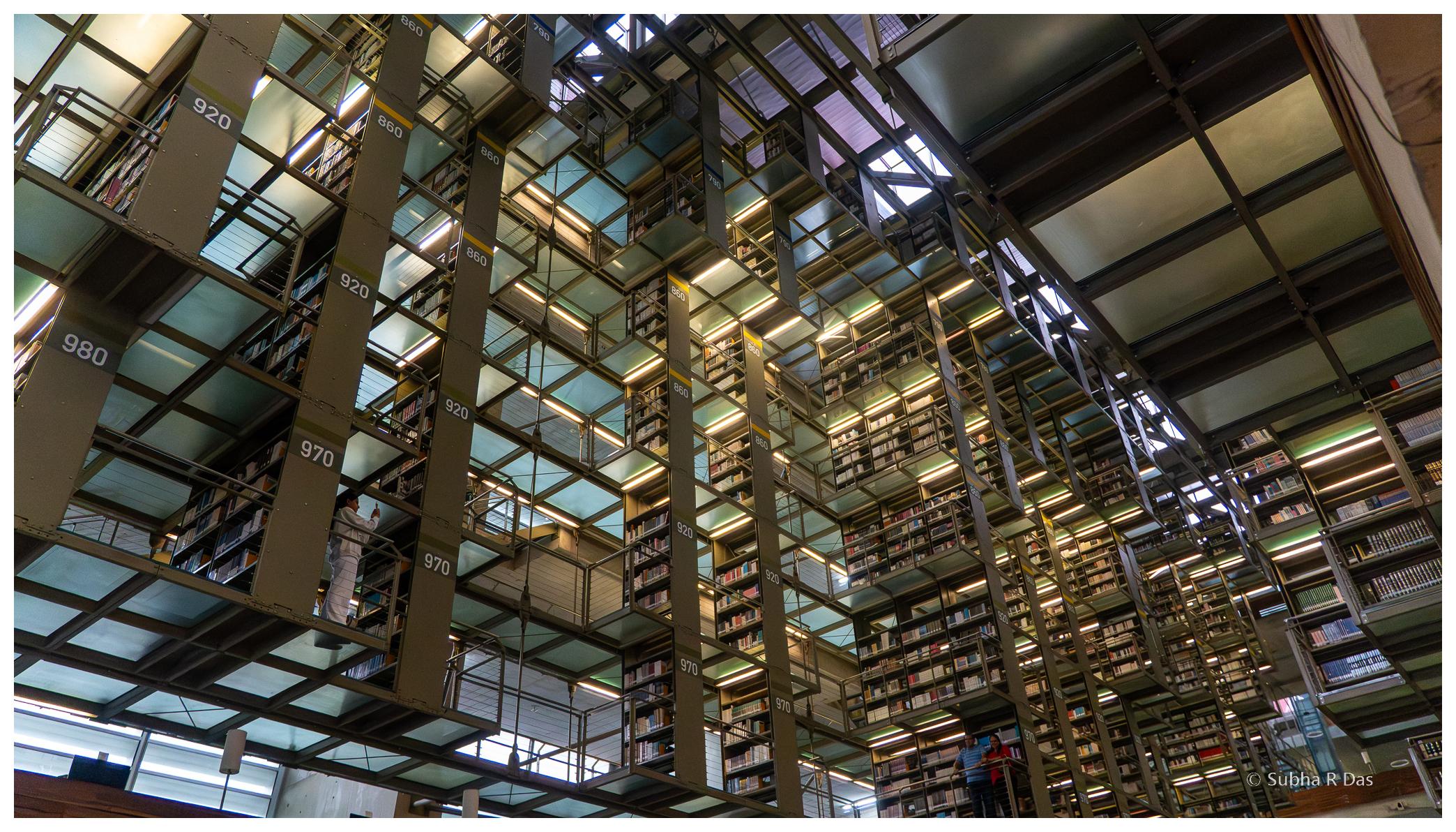 Looking up at the multi storied shelving and frosted glass floors in the library. A person (left bottom of frame) is taking a picture with their phone of two people further away (bottome, right of frame). Looking up at the multi storied shelving and frosted glass floors in the library. A person (left bottom of frame) is taking a picture with their phone of two people further away (bottome, right of frame).