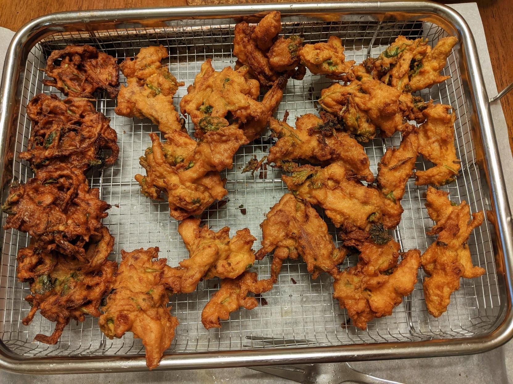 Metal mesh tray with a few onion pakodas to the left and a bunch of odd shaped pieces of brown fried battered shredded chicken