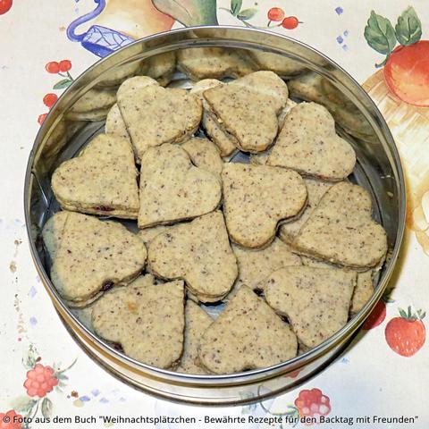 Auf dem Tisch steht eine Blechdose mit gefüllten Nussherzen. Die Textur der Kekse ist braun gesprenkelt wegen der gemahlenen Haselnüsse im Teig