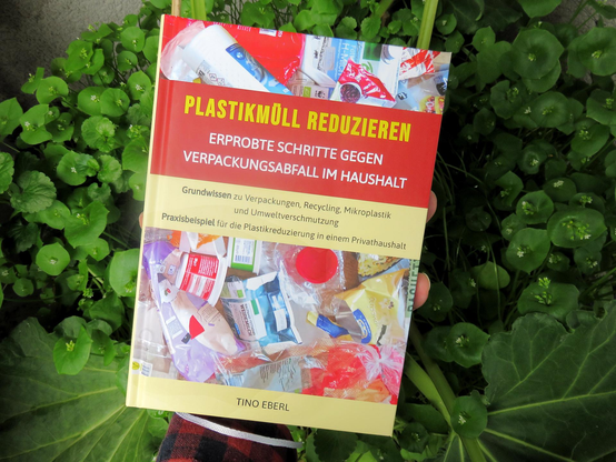 Eine Hand hält ein Buch mit einem Cover, das Bilder von Plastikverpackungen zeigt. Der obere Teil des Buchumschlags ist rot und der untere beige. Im Hintergrund befinden sich Rhabarberpflanzen und Winterportulak in einem Kübel in dichter Anordnung. 