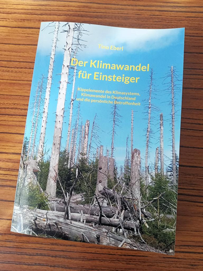 Buch mit einem Coverbild, das einen Wald mit vielen abgestorbenen, kahlen Bäumen zeigt. Die Baumstämme sind dünn und größtenteils ohne Äste, einige sind abgebrochen. Am Boden liegen mehrere umgestürzte Baumstämme. Dazwischen wachsen einige grüne, junge Nadelbäume. Der Himmel ist blau und wolkenlos. Das Buch liegt auf einer Holzoberfläche mit klaren, horizontal verlaufenden Maserungen.