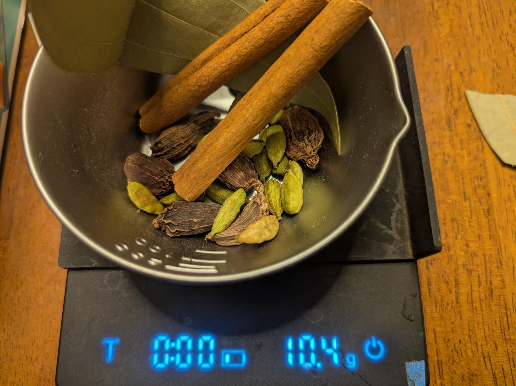 A small metal bowl on a digital scale. The bowl has black and green cardamom pods and 2 sticks of cinnamon and a bent bay leaf