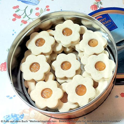 Auf dem Tisch mit Früchtetischdecke steht eine blau-gelbe Blechdose mit hübsch anzusehenden Schmalzblumen. Die Kekse in Blumenform haben in der Mitte ein Lock und die Marmelade scheint Orange durch, so dass es wie eine Blüte ausieht.