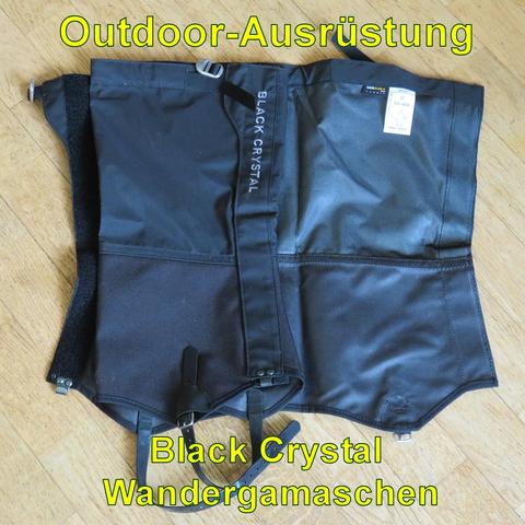 Ein Paar schwarze Wandergamaschen der Marke Black Crystal liegt flach auf einem Holzboden. Die Gamaschen bestehen aus robustem, wasserabweisendem Material und haben an den Seiten Klettverschlüsse zum einfachen Anlegen. Unten befinden sich verstellbare Riemen zur Befestigung unter dem Schuh. Ein kleiner Metallhaken ist an der Vorderseite zur Sicherung an den Schnürsenkeln angebracht. Die Gamaschen bieten Schutz vor Nässe und Schmutz, ideal für Wanderungen in anspruchsvollem Gelände.