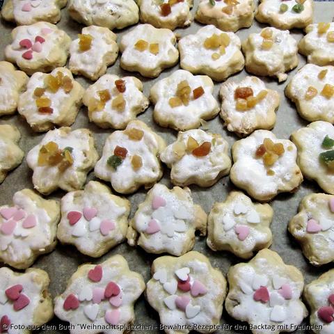 Auf Backpapier liegen Mürbeteigpplätzchen in Blumenform mit Zuckerguss und Zuckerherzchen. Manche haben kandierte Fruchtstücke in grün, gelb, rot und orange als Dekoration.