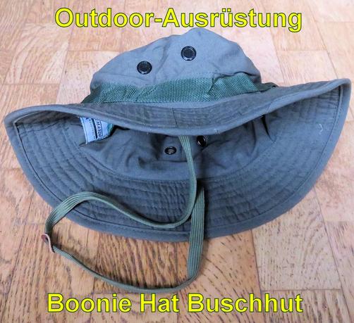 Ein olivgrüner Boonie Hat Buschhut liegt auf einem Holzboden. Der Hut hat eine breite, flexible Krempe und mehrere Lüftungsösen aus Metall an den Seiten. Ein grünes Band verläuft um den Hut für zusätzliche Befestigungsmöglichkeiten. An der Unterseite befindet sich ein verstellbares Kinnband, um den Hut sicher zu fixieren. Der robuste Stoff und das Design eignen sich ideal für Outdoor-Aktivitäten wie Wandern oder Camping, um Schutz vor Sonne und Regen zu bieten.