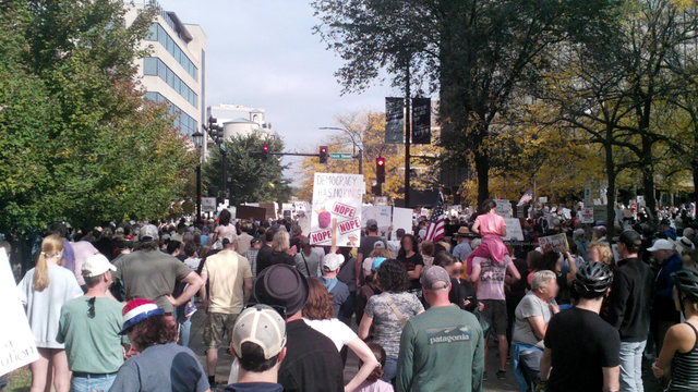 Looking North: A large crowd of people marching down a tree-lined city street during a protest, holding signs including one that reads "Democracy has no kings" and "NOPE."