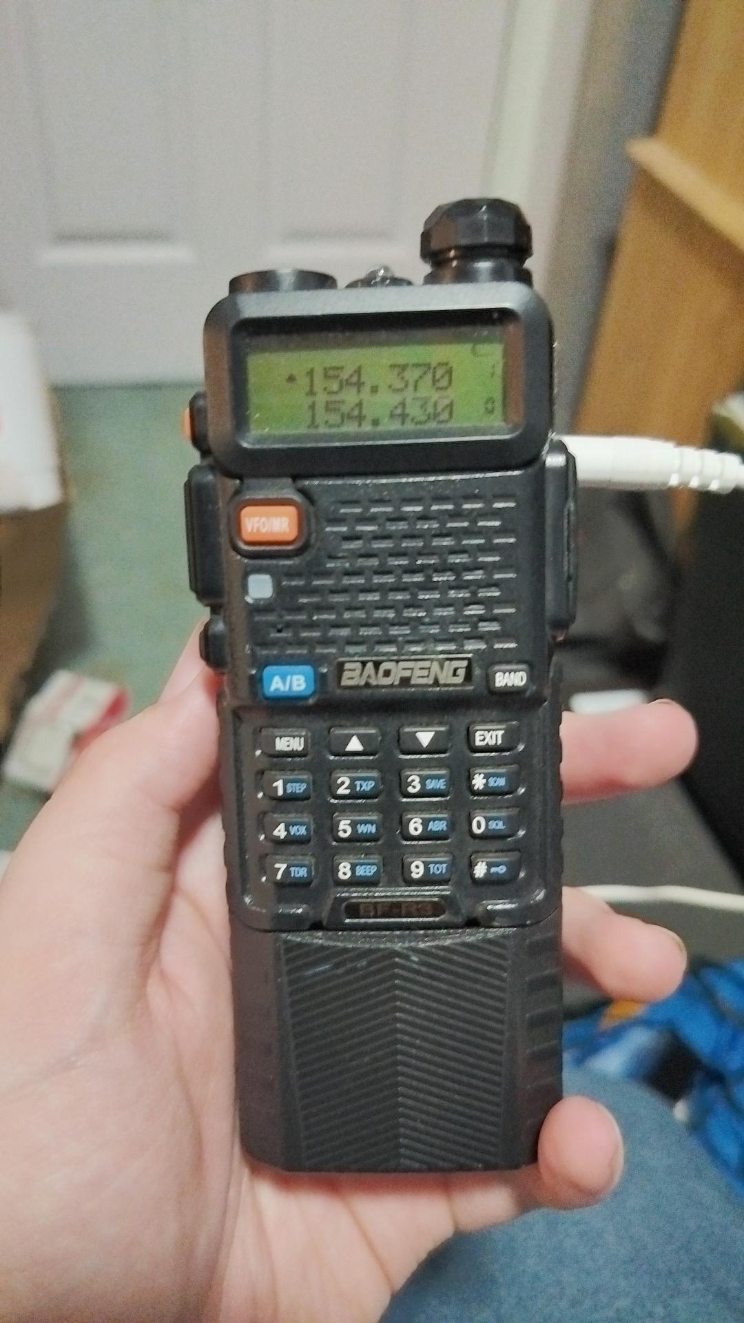 A black Baofeng BF-R3 handheld radio with green display showing 154.370 and 154.430 frequency values, held in a person's hand with white cable plugged in the side.