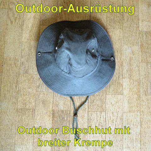 Ein olivgrüner Outdoor-Buschhut mit breiter Krempe liegt flach auf einem Holzboden. Der Hut hat mehrere Metallösen zur Belüftung sowie ein verstellbares Kinnband mit einem Kordelstopper, um ihn sicher zu fixieren. Das Material wirkt robust und wasserabweisend, ideal für Schutz vor Sonne und Regen bei Outdoor-Aktivitäten. Die breite Krempe bietet zusätzlichen Sonnenschutz.