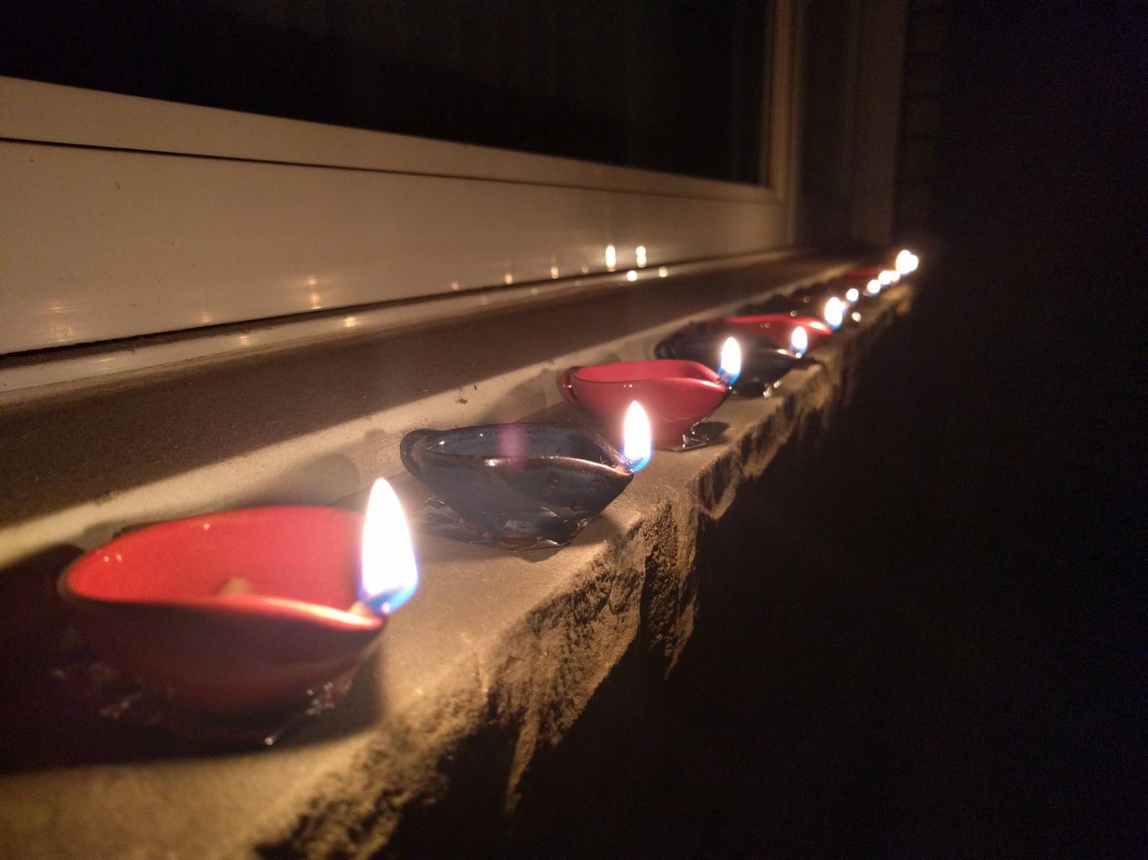 A picture of small ceramic oil lamps, red and blue with small cotton wicks and flames, all in a row in a narrow windowsill outside in the dark