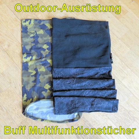 Mehrere Buff Multifunktionstücher liegen auf einem Holzboden. Eines hat ein Tarnmuster in Grün- und Brauntönen, während die anderen dunkler und teils mit Paisley-Muster verziert sind. Die Tücher sind teilweise gefaltet und bestehen aus verschiedenen Materialien, darunter ein schwarzes, fleeceartiges Tuch. Sie sind vielseitig einsetzbar, zum Beispiel als Halstuch, Stirnband oder Schal, ideal für Outdoor-Aktivitäten.