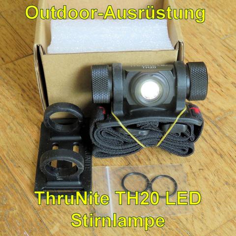 Die ThruNite TH20 LED Stirnlampe ist auf einem Holzboden zusammen mit Zubehör abgebildet. Die Lampe hat ein kompaktes, schwarzes Design mit gerändelten Seiten für besseren Halt. Sie ist auf einem elastischen Stirnband befestigt, das zusammengerollt und mit einem Gummiband gesichert ist. Neben der Lampe befinden sich zwei Ersatzdichtungen und eine zusätzliche Halterung. Im Hintergrund steht eine offene Pappverpackung mit Schaumstoffeinlage. Die Stirnlampe wirkt robust und ist für Outdoor-Aktivit…