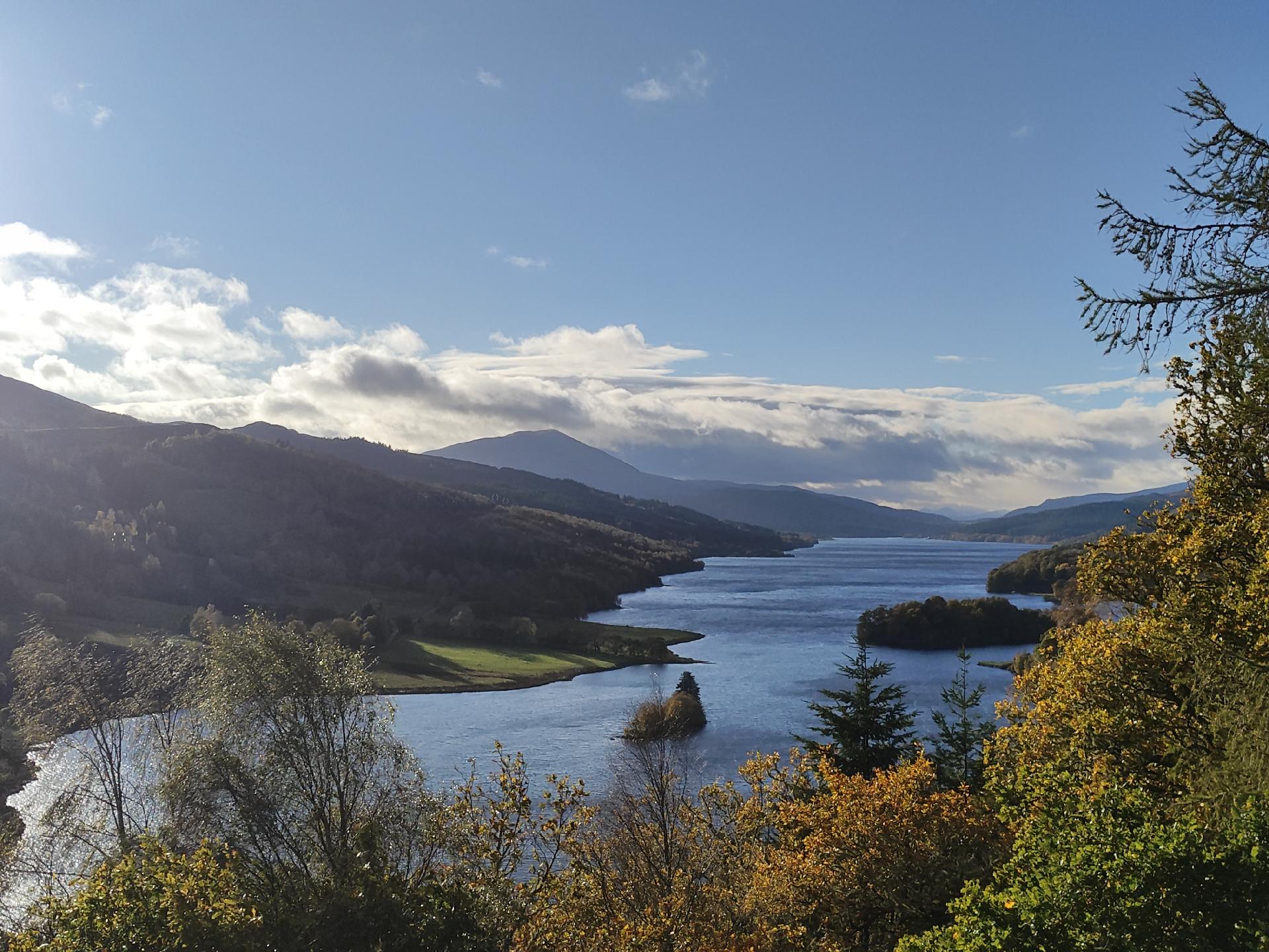 View from high up along a loch looking west. Autumn trees all around. Blue sky.
