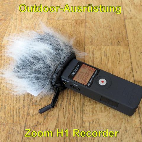 Ein Zoom H1 Rekorder liegt auf einem Holzboden. Der kompakte, schwarze Audiorekorder hat ein kleines Display und Bedientasten an der Seite. Oben befindet sich ein Mikrofon mit einem großen, flauschigen Windschutz in Grau und Weiß, um Windgeräusche bei Außenaufnahmen zu minimieren. Der Rekorder verfügt über eine Aufnahme-Taste sowie Anschlüsse für Kopfhörer und Line-Out. Er ist ideal für Outdoor-Audioaufnahmen, Interviews oder Naturgeräusche.