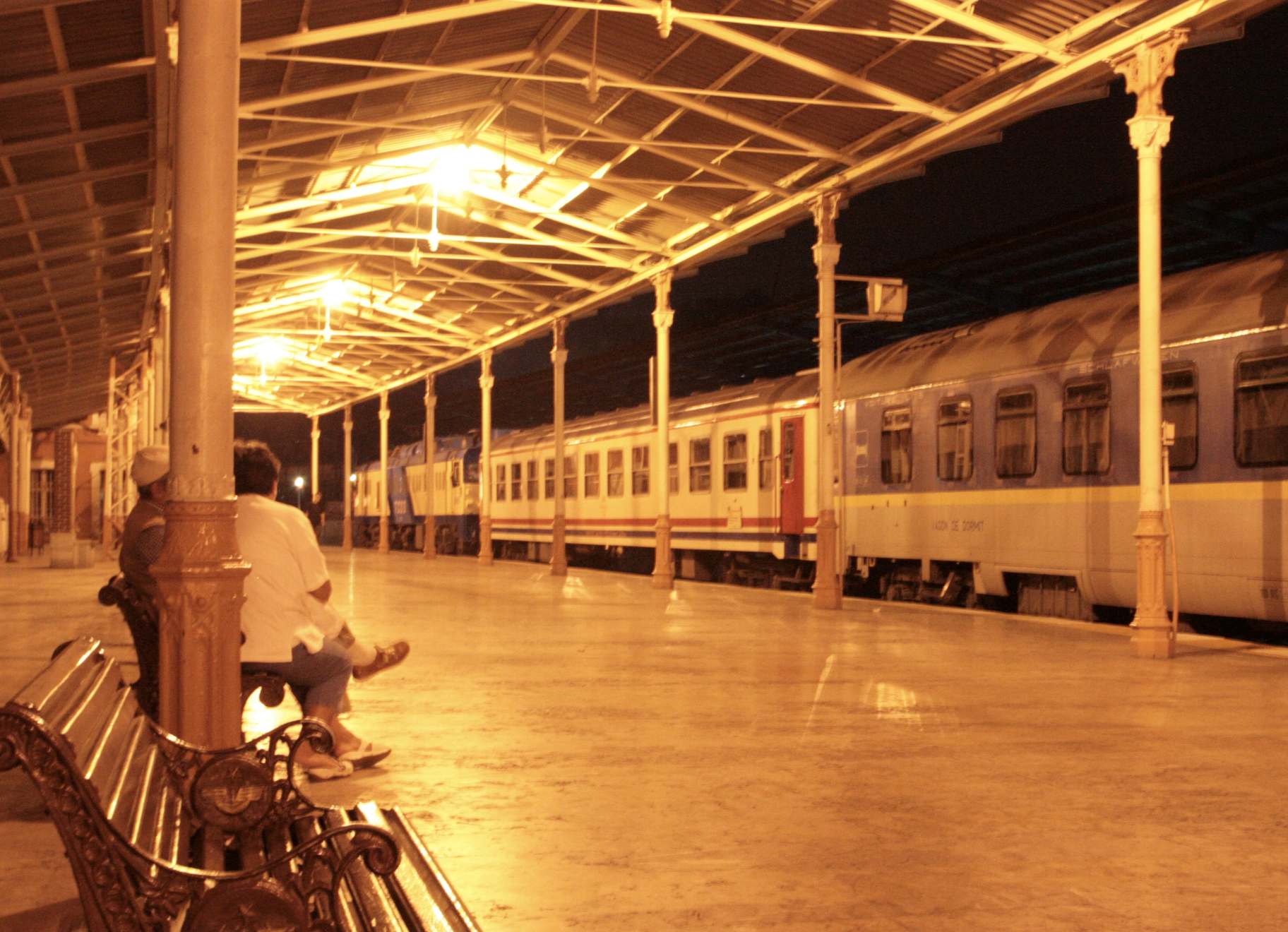 starting in the Istanbul train station, pictured at night in yellow lighting, a train waiting on the tracks near the smooth stone platform, which has a roof over it. a few benches sit to the left